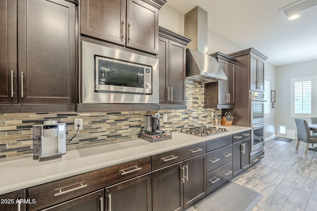 kitchen featuring dark brown cabinetry, stainless steel appliances, light countertops, and wall chimney range hood