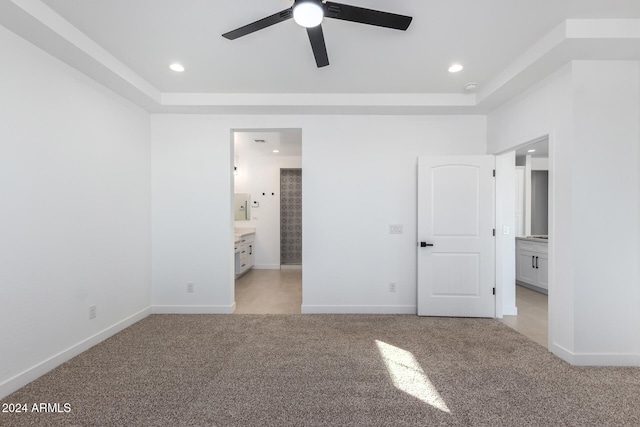 unfurnished bedroom with a tray ceiling, light colored carpet, ceiling fan, and ensuite bath