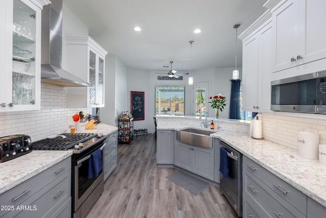 kitchen with pendant lighting, glass insert cabinets, appliances with stainless steel finishes, a sink, and wall chimney exhaust hood