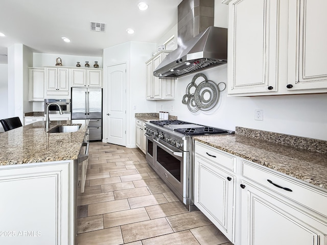 kitchen featuring stone counters, high end appliances, visible vents, a sink, and wall chimney range hood