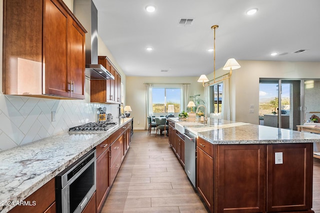 kitchen featuring sink, wall chimney exhaust hood, an island with sink, decorative light fixtures, and stainless steel appliances