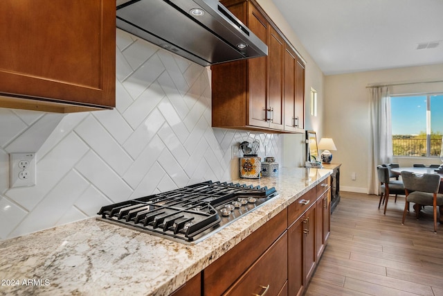 kitchen with light wood-type flooring, backsplash, light stone counters, stainless steel gas cooktop, and range hood