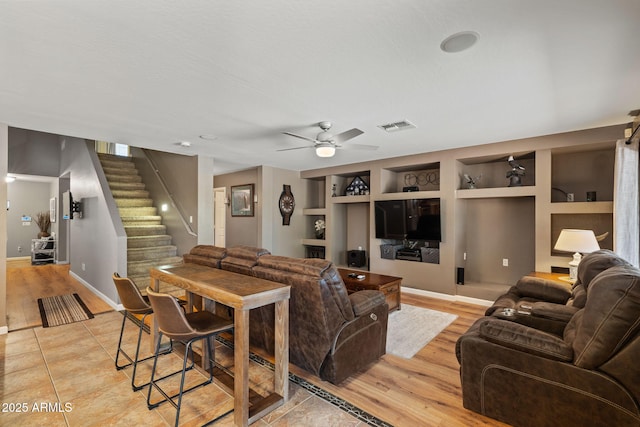 living room with built in shelves, ceiling fan, stairway, baseboards, and light wood-style floors