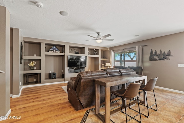 living room featuring ceiling fan, built in shelves, baseboards, a textured ceiling, and light wood-type flooring