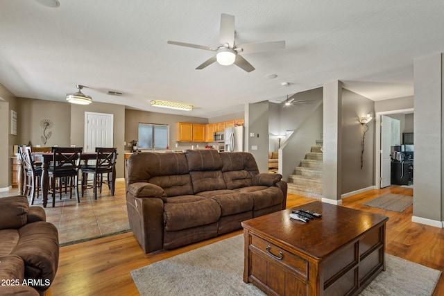 living area with a ceiling fan, light wood-style flooring, stairs, and baseboards