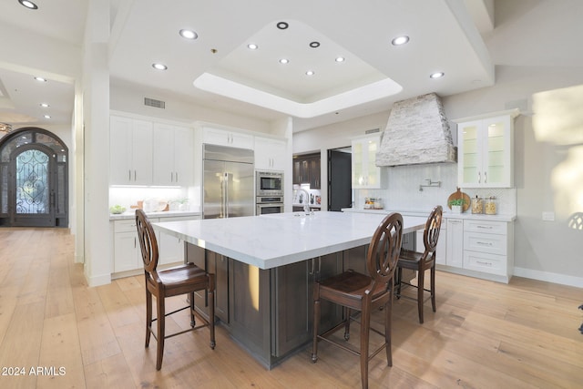 kitchen with a kitchen bar, built in appliances, a large island with sink, custom range hood, and white cabinets