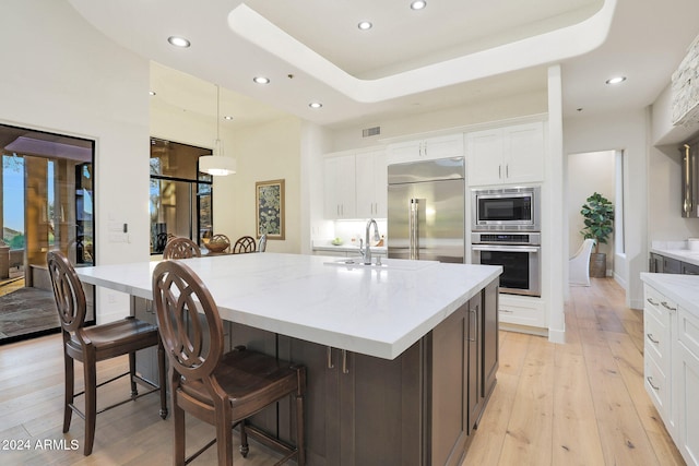 kitchen featuring built in appliances, a raised ceiling, pendant lighting, a kitchen island with sink, and white cabinets