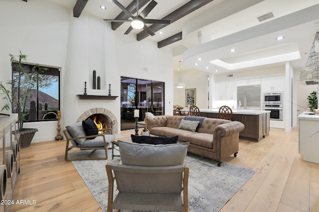 living room with beamed ceiling, a large fireplace, a high ceiling, ceiling fan, and light hardwood / wood-style flooring