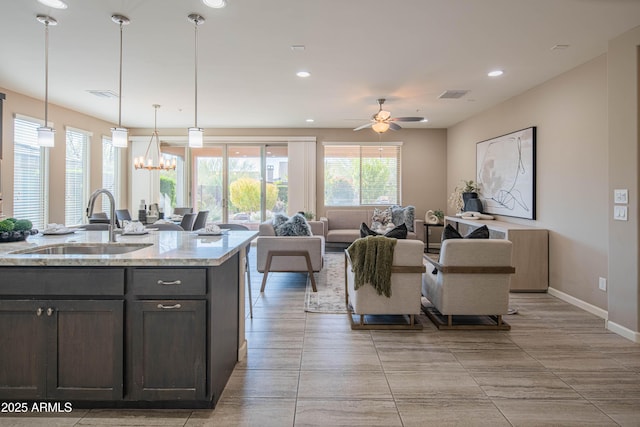 kitchen with dark brown cabinets, hanging light fixtures, light stone countertops, ceiling fan with notable chandelier, and sink