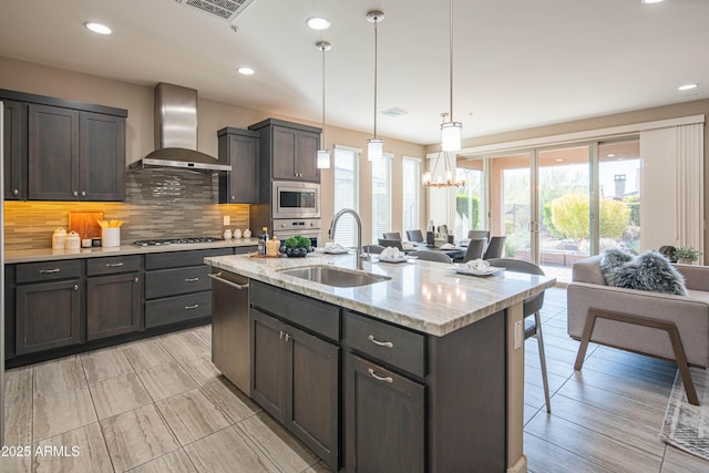 kitchen featuring backsplash, a kitchen island with sink, wall chimney range hood, pendant lighting, and light stone counters