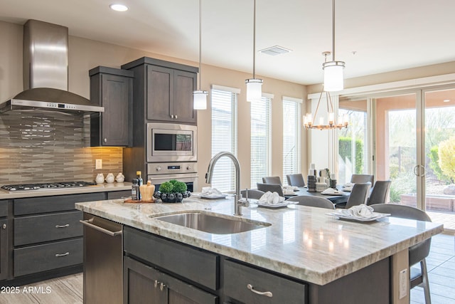 kitchen featuring a center island with sink, appliances with stainless steel finishes, light stone countertops, wall chimney exhaust hood, and sink