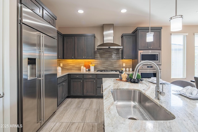 kitchen featuring light stone countertops, decorative light fixtures, wall chimney range hood, built in appliances, and sink