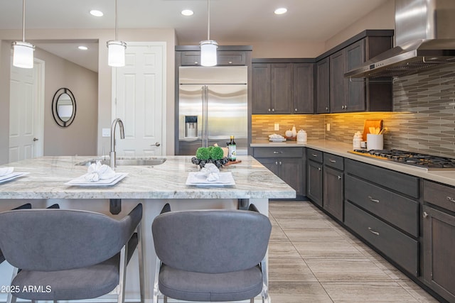 kitchen featuring hanging light fixtures, appliances with stainless steel finishes, a center island with sink, and wall chimney exhaust hood