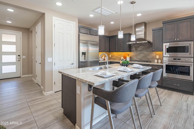 kitchen with wall chimney exhaust hood, built in appliances, sink, a kitchen island with sink, and a breakfast bar