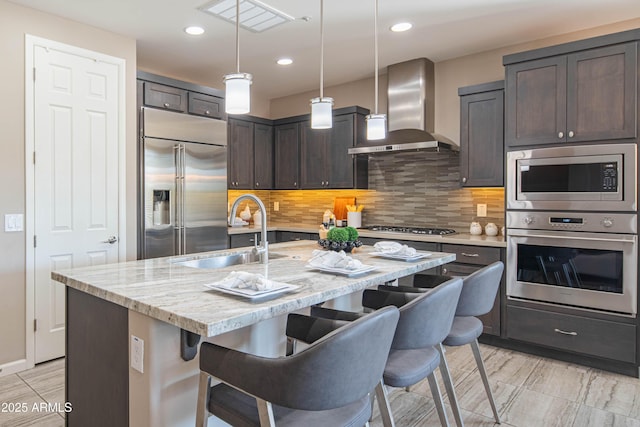 kitchen with decorative light fixtures, sink, built in appliances, an island with sink, and wall chimney exhaust hood