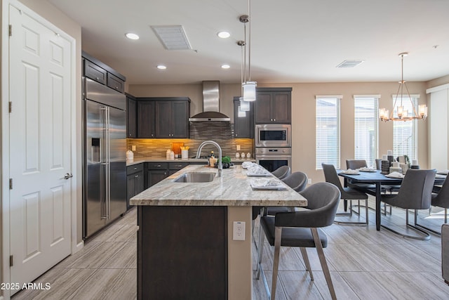 kitchen featuring wall chimney exhaust hood, built in appliances, tasteful backsplash, sink, and a center island with sink