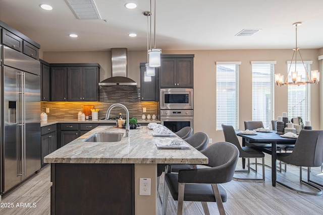 kitchen with decorative light fixtures, wall chimney range hood, built in appliances, an island with sink, and backsplash