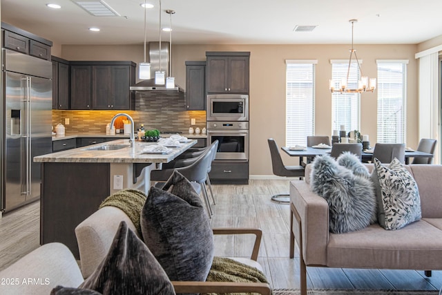 kitchen featuring backsplash, a kitchen island with sink, built in appliances, wall chimney exhaust hood, and sink