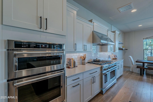 kitchen featuring backsplash, white cabinets, wall chimney range hood, light wood-type flooring, and stainless steel appliances