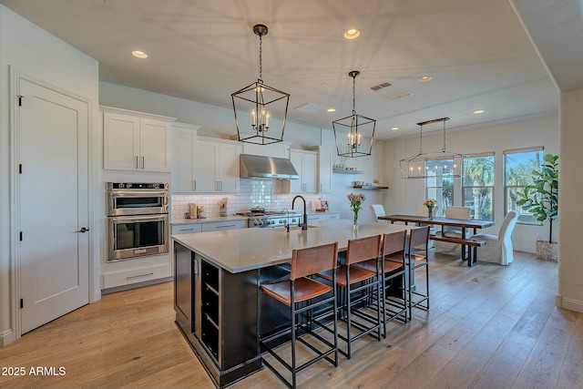 kitchen with an island with sink, white cabinetry, double oven, and wall chimney range hood