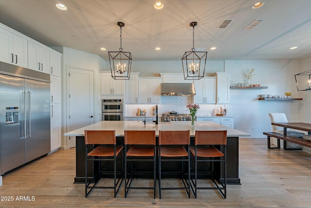 kitchen with a kitchen island with sink, wall chimney exhaust hood, appliances with stainless steel finishes, decorative light fixtures, and white cabinetry