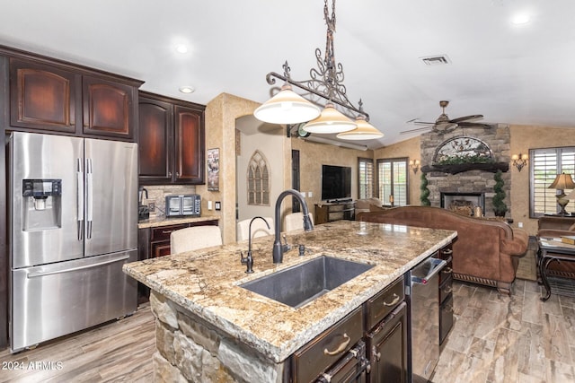 kitchen featuring vaulted ceiling, sink, stainless steel fridge with ice dispenser, a stone fireplace, and an island with sink