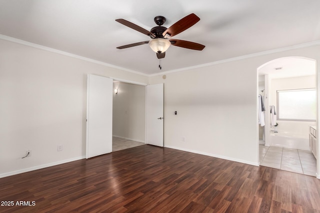 empty room with arched walkways, a ceiling fan, wood finished floors, ornamental molding, and baseboards