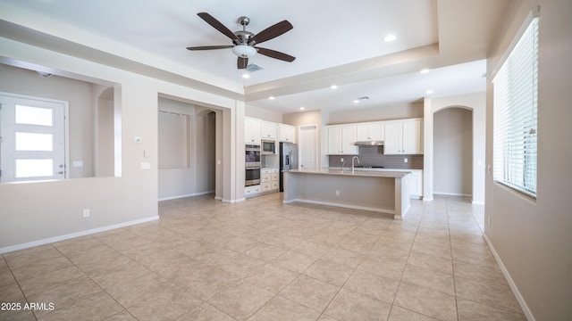 kitchen featuring decorative backsplash, ceiling fan, a center island with sink, arched walkways, and plenty of natural light