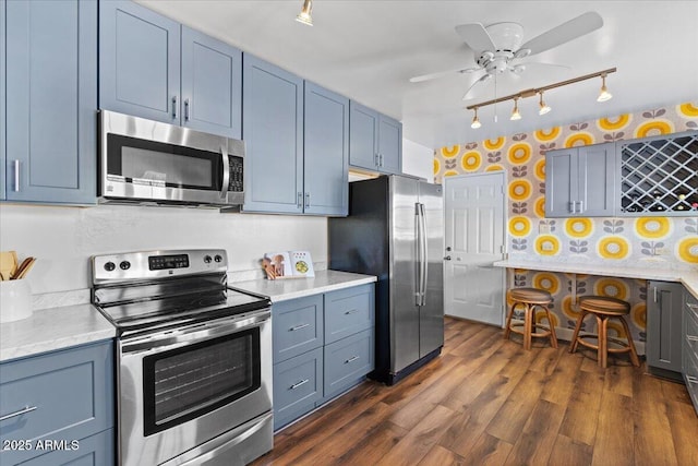 kitchen with blue cabinetry, rail lighting, ceiling fan, appliances with stainless steel finishes, and dark hardwood / wood-style floors