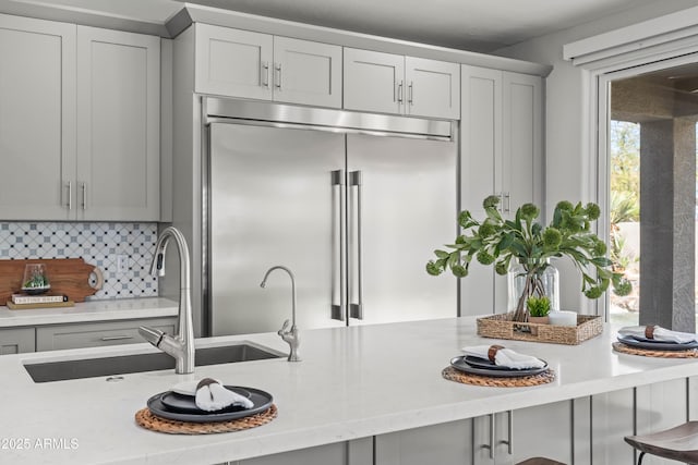 kitchen featuring gray cabinetry, built in refrigerator, a sink, decorative backsplash, and a kitchen bar