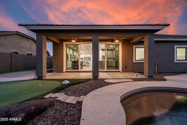 back of house at dusk with a ceiling fan, a patio area, fence, and stucco siding