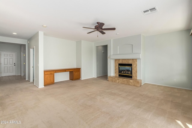 unfurnished living room with a tile fireplace, built in desk, light colored carpet, and ceiling fan