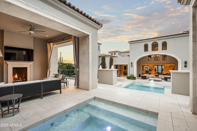 pool at dusk featuring ceiling fan, a jacuzzi, a patio area, and an outdoor living space with a fireplace