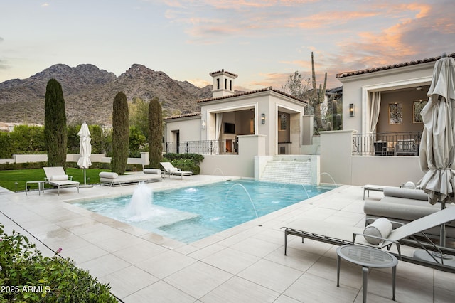 pool at dusk with pool water feature, a mountain view, and a patio area