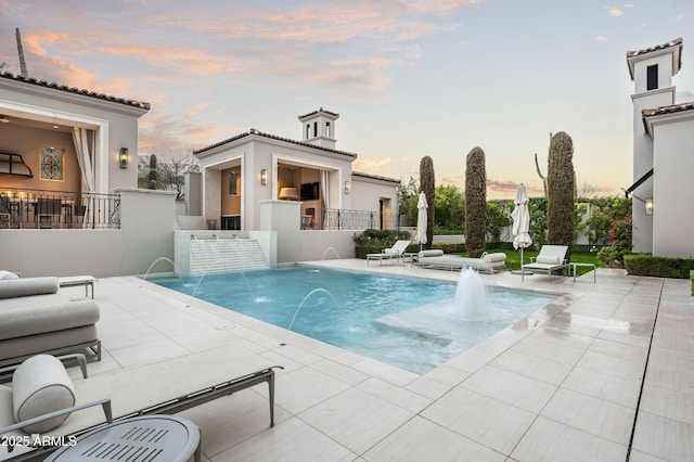 pool at dusk featuring pool water feature, a fireplace, and a patio area