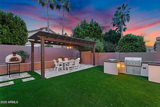 yard at dusk with exterior kitchen, a fenced backyard, a pergola, and a patio area