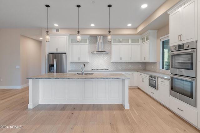 kitchen with white cabinets, appliances with stainless steel finishes, an island with sink, and wall chimney range hood