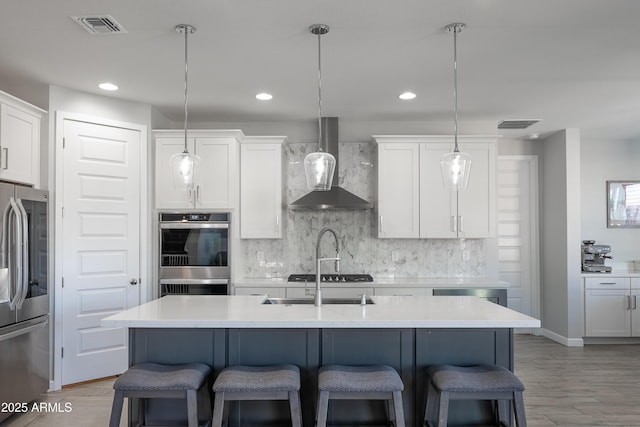 kitchen featuring white cabinetry, wall chimney range hood, stainless steel appliances, an island with sink, and hanging light fixtures