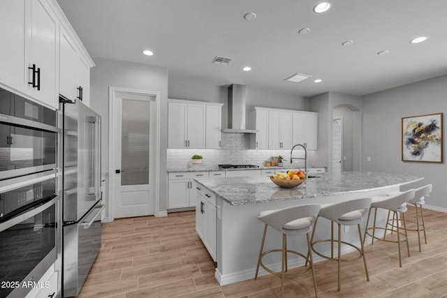 kitchen with white cabinetry, wall chimney range hood, an island with sink, and appliances with stainless steel finishes