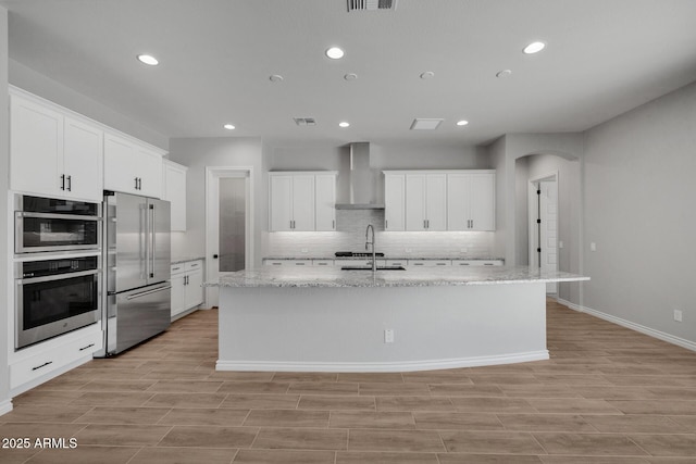 kitchen featuring a kitchen island with sink, wall chimney range hood, white cabinetry, and appliances with stainless steel finishes