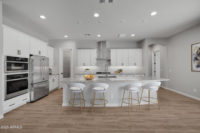 kitchen featuring white cabinets, appliances with stainless steel finishes, an island with sink, and wall chimney range hood