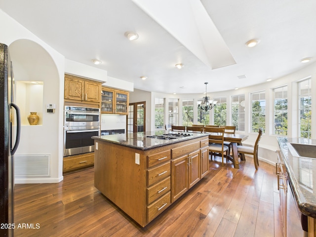 kitchen featuring brown cabinetry, stainless steel appliances, a kitchen island with sink, dark wood-style flooring, and dark stone countertops