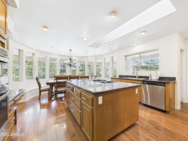 kitchen featuring stainless steel appliances, a sink, brown cabinetry, a chandelier, and dark stone countertops