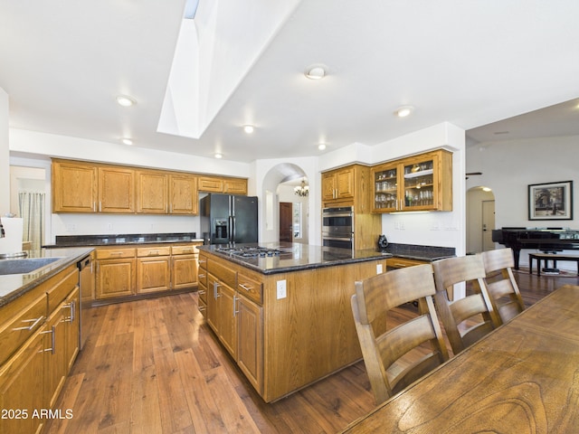 kitchen with arched walkways, appliances with stainless steel finishes, brown cabinets, hardwood / wood-style floors, and a kitchen island