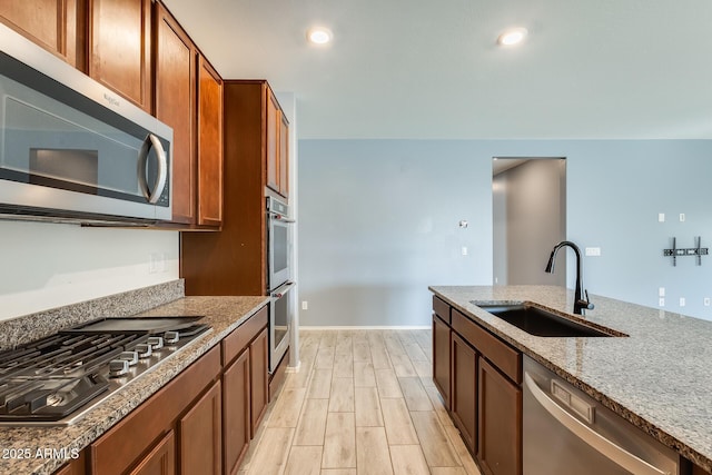 kitchen with brown cabinetry, appliances with stainless steel finishes, light stone countertops, wood finish floors, and a sink