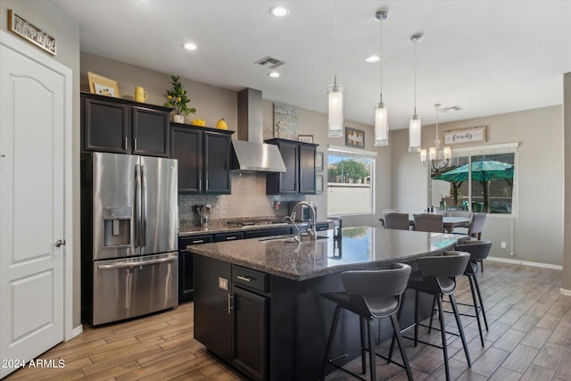 kitchen with hanging light fixtures, stainless steel refrigerator with ice dispenser, tasteful backsplash, wall chimney exhaust hood, and a kitchen island with sink