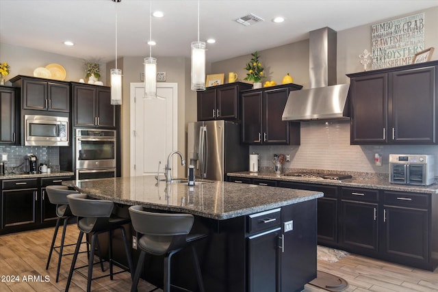 kitchen with an island with sink, appliances with stainless steel finishes, wall chimney range hood, and tasteful backsplash