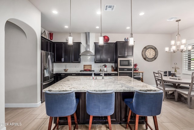 kitchen featuring wall chimney range hood, light hardwood / wood-style flooring, pendant lighting, a kitchen island with sink, and appliances with stainless steel finishes