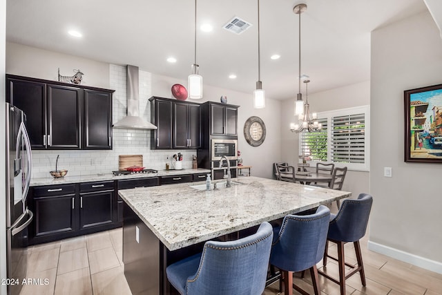 kitchen featuring wall chimney exhaust hood, stainless steel appliances, a kitchen island with sink, sink, and decorative light fixtures