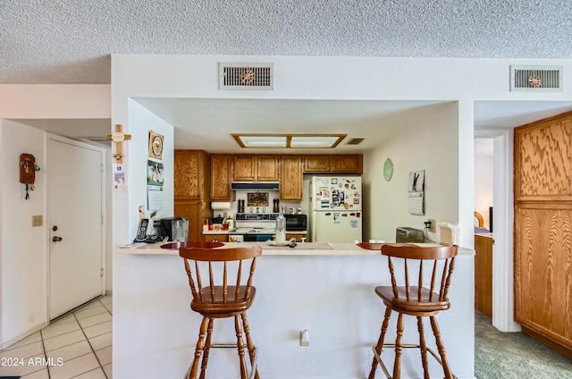 kitchen featuring kitchen peninsula, a textured ceiling, white appliances, a kitchen bar, and light tile patterned floors